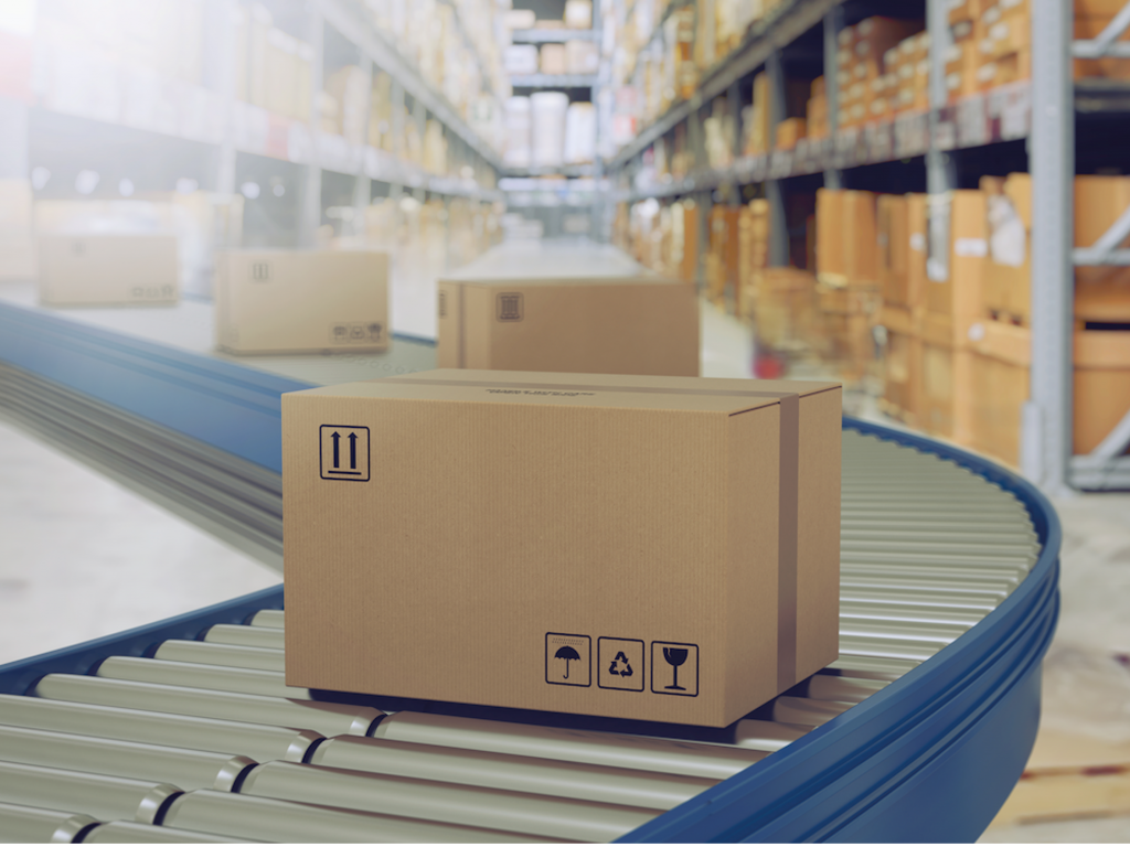 Cardboard boxes traveling down a conveyor system, with racks of boxes in the background